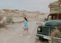 Vista laterale della donna in pantaloncini e camicia di jeans che cammina in camion verde e guarda lontano nella natura — Foto stock