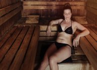 Frau im schwarzen Bikini sitzt mit geschlossenen Augen auf Handtuch im Dampfbad, lehnt an Holzbank und genießt Hitze — Stockfoto