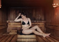 Femme en bikini noir assise avec les yeux fermés sur une serviette dans un hammam appuyée sur un banc en bois et bénéficiant de la chaleur — Photo de stock