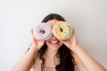 Безтурботна жінка розважається і грає з глазурованими пончиками зі зморшками, стоячи біля білої стіни — стокове фото