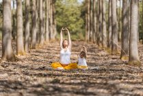 Беременная женщина и маленькая девочка с закрытыми глазами в белых рубашках и желтых брюках растягиваются, сидя на земле и практикуя йогу на поляне в лесу — стоковое фото