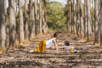 Вид сбоку беременной женщины и маленькой девочки, растянувшейся на земле во время занятий йогой в лесу — стоковое фото
