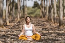 A piedi nudi serena donna incinta con gli occhi chiusi seduta in posa padmasana e meditando nella foresta — Foto stock