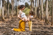 Side view of cheerful pregnant woman and little girl touching bellies in autumn forest glade — Stock Photo