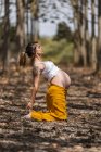 Side view of pregnant female with closed eyes in white shirt and yellow pants standing on knees while yoga in glade in forest — Stock Photo