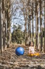 Взрослая безмятежная женщина медитирует с голубым мячом, сидя на земле среди деревьев в парке — стоковое фото
