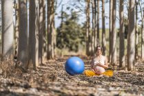 Calm pregnant lady with closed eyes touching belly while sitting and meditating beside big blue pilates fit ball in forest glade — Stock Photo
