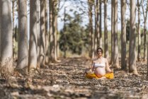 Calma mujer embarazada adulta meditando mientras está sentado en pose de loto en el suelo en el parque - foto de stock