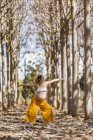 Взрослая беременная женщина в белой рубашке и желтых брюках стоит с протянутыми руками и практикует йогу среди деревьев — стоковое фото