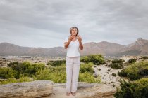 Frau hält die Hände über dem Kopf zusammen und schließt die Augen, während sie in der Natur auf einem Felsen steht und meditiert — Stockfoto