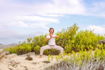 Женщина стоит на песке возле зеленого кустарника и медитирует, практикуя Тай Чи против облачного неба в природе — стоковое фото
