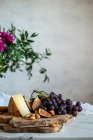 Figos maduros fatiados e uvas azuis escuras ao lado de pedaço de queijo em tábuas de corte de madeira perto do buquê de flores rosa entre folhas verdes contra a parede cinza borrada — Fotografia de Stock