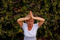 Von oben von einer Frau mit geschlossenen Augen und gefalteten Händen, die neben grünem Strauch steht und in der Landschaft meditiert — Stockfoto