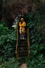Длинноволосая женщина в жёлтой куртке и английская указка на деревянной лестнице в зелёном заборе растения в дождливую погоду — стоковое фото