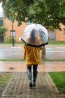 Rückansicht einer Frau in gelbem Regenmantel und dunklen Gummistiefeln mit transparentem Regenschirm, die unter Regen auf der Straße steht — Stockfoto