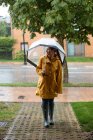 Giovane donna in impermeabile giallo e stivali di gomma scura con ombrello trasparente in piedi sotto la pioggia in strada e guardando altrove — Foto stock