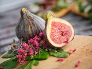 Ripe whole and sliced figs among flavored green leaves of herbs and colorful flowers on wooden cutting board — Stock Photo