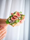 Person hält hausgemachtes offenes Sandwich mit Feigen- und Käsescheiben auf Roggenbrot mit Rucola — Stockfoto