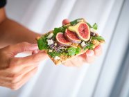 Person holding homemade open sandwich with slices of fig and cheese on rye bread with rocket salad — Stock Photo