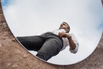 Vista de ángulo bajo de la reflexión del hombre de ensueño en camisa y tirantes de pie sobre el cielo azul en espejo oval en suelo polvoriento - foto de stock