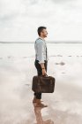 Вид сбоку на человека в белой рубашке и подтяжки с потрепанным портфелем, стоящего босиком на мрачном пляже — стоковое фото