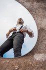 Reflexão de baixo ângulo do homem sonhador na camisa e suspensórios em pé sobre o céu azul no espelho oval no chão empoeirado — Fotografia de Stock