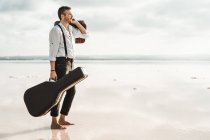 Side view of serious man in white shirt and suspenders carrying guitar and briefcase while standing barefoot in water by shore — Stock Photo