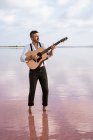 Страстный мужчина в белой рубашке и подтяжках, играющий на гитаре, стоя босиком в воде на берегу — стоковое фото