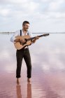 Leidenschaftlicher Mann in weißem Hemd und Hosenträgern, der am Ufer barfuß im Wasser steht und Gitarre spielt — Stockfoto