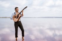 Passionate man in white shirt and suspenders playing guitar while standing barefoot in water by shore — Stock Photo