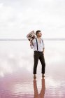 Passionate man in white shirt and suspenders holding guitar while standing barefoot in water by shore on cloudy weather — Stock Photo