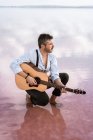Wistful man in white shirt and susenders playing acoustic guitar and looking away surrounded with smooth sea water — стоковое фото