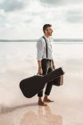 Вид збоку серйозного чоловіка в білій сорочці і підтяжки з гітарою і портфелем, стоячи босоніж у воді біля берега — стокове фото