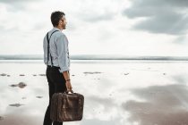 Side view of man carrying shabby briefcase while standing barefoot looking away on gloomy beach — Stock Photo