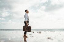 Side view of man carrying shabby briefcase while standing barefoot looking away on gloomy beach — Stock Photo