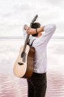 From behind man in white shirt holding acoustic guitar behind back while standing on shore on cloudy weather — Stock Photo