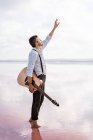 Melancholic passionate man in white shirt and suspenders holding guitar while standing barefoot in water raising hand to sky — Stock Photo