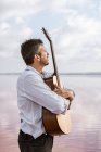 Melancholic passionate man in white shirt and suspenders embracing guitar while standing in water by shore — Stock Photo