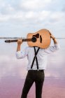 Da dietro l'uomo in camicia bianca che tiene la chitarra acustica dietro la schiena mentre in piedi sulla riva sul tempo nuvoloso negli Stati Uniti — Foto stock