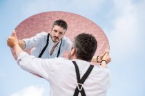 Low angle reflection of pensive man in shirt and suspenders standing over blue sky and lifting oval mirror — Stock Photo