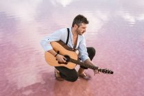 Сміливий чоловік грає на акустичній гітарі, сидячи на пляжі в оточенні гладкого моря, що відображає величний хмарний пейзаж — стокове фото