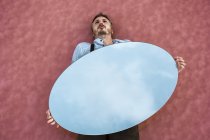 From above pensive man lying down on pink water holding oval mirror reflecting blue sky — Stock Photo