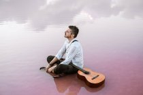 Uomo malinconico con chitarra acustica seduto sulla spiaggia guardando lontano circondato da un mare liscio che riflette maestoso paesaggio nuvoloso — Foto stock