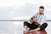 Uomo malinconico che suona la chitarra acustica seduto sulla spiaggia circondato da un mare liscio che riflette maestoso paesaggio nuvoloso — Foto stock