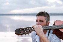 Man in white shirt and suspenders carrying acoustic guitar and sitting on beach by water — Stock Photo