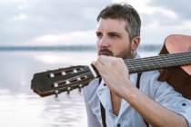 Чоловік у білій сорочці і підтяжки, що несуть акустичну гітару і сидять на пляжі біля води — стокове фото