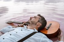 Зверху чоловік у білій сорочці і підтяжки лежать на плаваючій акустичній гітарі в морі на піщаному березі — стокове фото