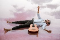 Человек с гитарой лежит на спине, глядя в сторону и протягивая руку к небу в море на песчаном берегу — стоковое фото