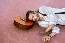 Сплячий чоловік лежить з закритими очима з акустичною гітарою в морі на піщаному березі — стокове фото