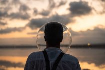 Back view of unrecognizable man in wet shirt with empty aquarium on head standing and contemplating by still sea in twilight — Stock Photo